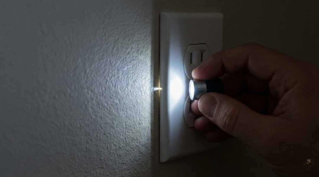 A person using a flashlight in a dark room to detect light leaking through a gap around an electrical outlet on a shared wall, indicating a sound leak pathway.